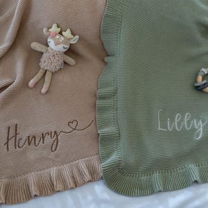 May include: Two knitted baby blankets, one tan and one sage green, with ruffled edges. The tan blanket has the name "Henry" embroidered, and a deer plush toy rests on it. The green blanket has the name "Lilly" embroidered, and a wooden teether.