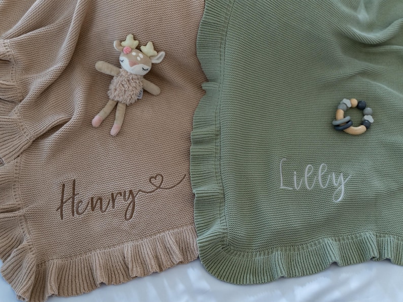 May include: Two knitted baby blankets, one tan and one sage green, with ruffled edges. The tan blanket has the name "Henry" embroidered, and a deer plush toy rests on it. The green blanket has the name "Lilly" embroidered, and a wooden teether.