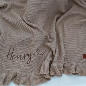 May include: A tan knitted baby blanket with a ruffled edge. The blanket has the name "Henry" embroidered in a cursive font, with a heart symbol. A small brown leather tag is sewn on the blanket.
