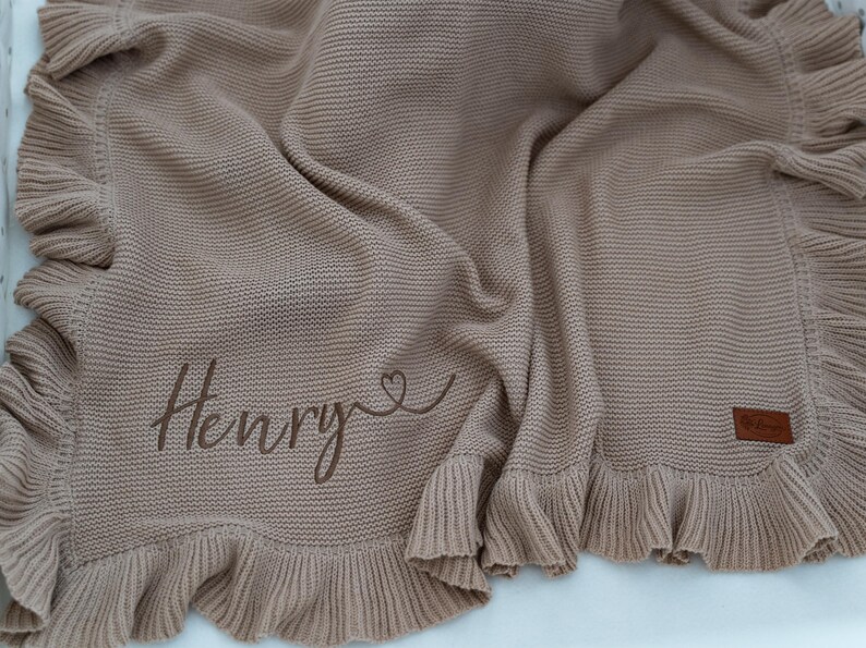May include: A tan knitted baby blanket with a ruffled edge. The blanket has the name "Henry" embroidered in a cursive font, with a heart symbol. A small brown leather tag is sewn on the blanket.
