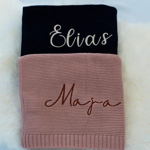 May include: Two personalized knitted blankets. The top blanket is navy blue with the name "Elias" embroidered in white script. The bottom blanket is dusty rose with the name "Maja" embroidered in brown script. Both blankets are folded.
