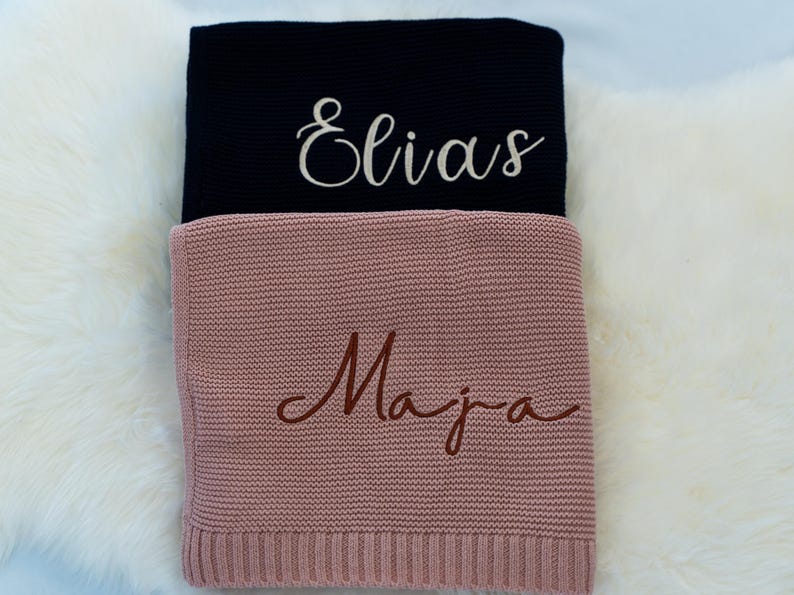 May include: Two personalized knitted blankets. The top blanket is navy blue with the name "Elias" embroidered in white script. The bottom blanket is dusty rose with the name "Maja" embroidered in brown script. Both blankets are folded.