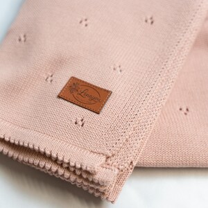 May include: A soft, dusty pink knitted blanket featuring a delicate pointelle pattern of small, spaced-out holes. A rectangular brown faux leather patch is stitched onto the fabric, displaying the embossed text Lenayou in a cursive script.