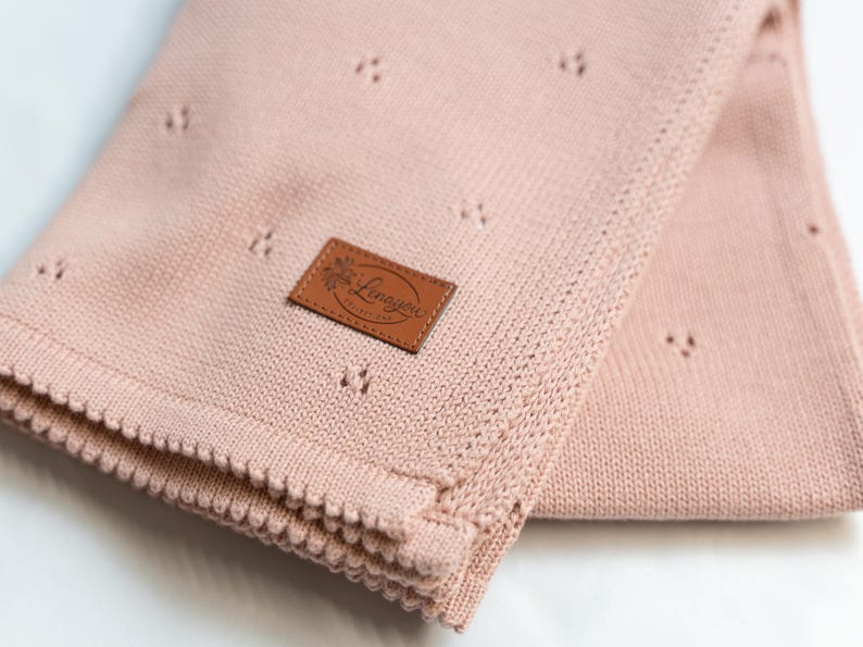 May include: A soft, dusty pink knitted blanket featuring a delicate pointelle pattern of small, spaced-out holes. A rectangular brown faux leather patch is stitched onto the fabric, displaying the embossed text Lenayou in a cursive script.