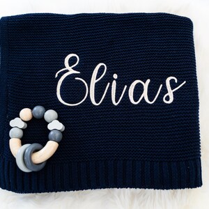 May include: Navy blue knitted baby blanket with the name "Elias" embroidered in white script. A wooden and silicone baby teether in shades of gray and natural wood is placed on the blanket.