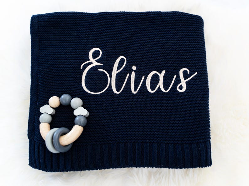 May include: Navy blue knitted baby blanket with the name "Elias" embroidered in white script. A wooden and silicone baby teether in shades of gray and natural wood is placed on the blanket.