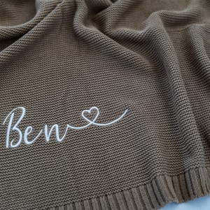 May include: A brown knitted blanket with the name "Ben" embroidered in white script, followed by a heart symbol. The blanket has a textured appearance and a ribbed border.