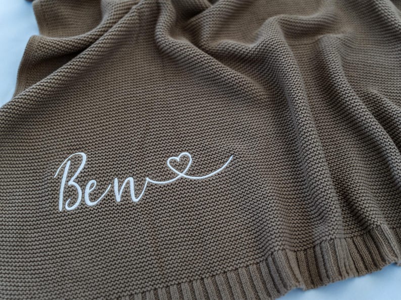 May include: A brown knitted blanket with the name "Ben" embroidered in white script, followed by a heart symbol. The blanket has a textured appearance and a ribbed border.