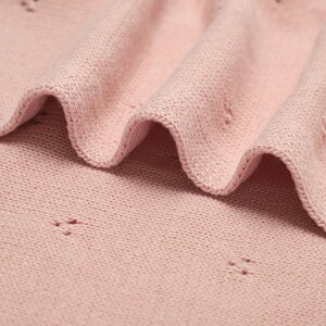 May include: A soft, light pink knitted fabric featuring a delicate pointelle pattern with small, evenly spaced eyelet clusters. The material is folded into gentle, flowing waves, highlighting the fine texture of the knit and the consistent pastel color throughout the textile surface.