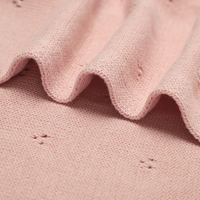 May include: A soft, light pink knitted fabric featuring a delicate pointelle pattern with small, evenly spaced eyelet clusters. The material is folded into gentle, flowing waves, highlighting the fine texture of the knit and the consistent pastel color throughout the textile surface.