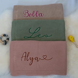 May include: Three personalized baby blankets in shades of tan, green, and pink, each embroidered with a name in a cursive font. A wooden teething ring and a yellow maraca are placed near the blankets.