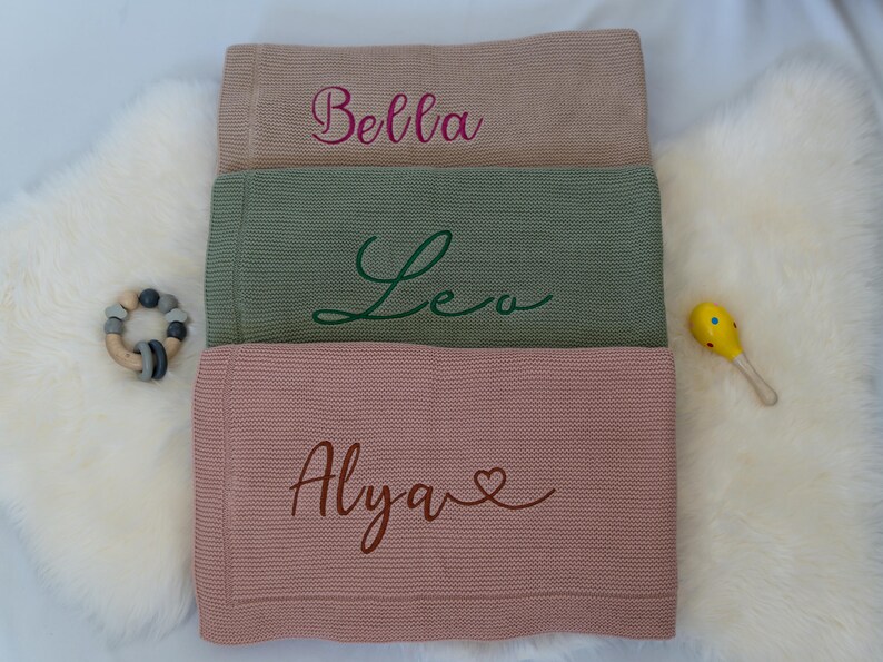 May include: Three personalized baby blankets in shades of tan, green, and pink, each embroidered with a name in a cursive font. A wooden teething ring and a yellow maraca are placed near the blankets.