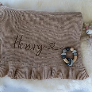 May include: A tan knitted baby blanket with a ruffled edge, embroidered with the name "Henry" and a heart. A wooden and silicone teether and a stuffed deer toy are next to the blanket.