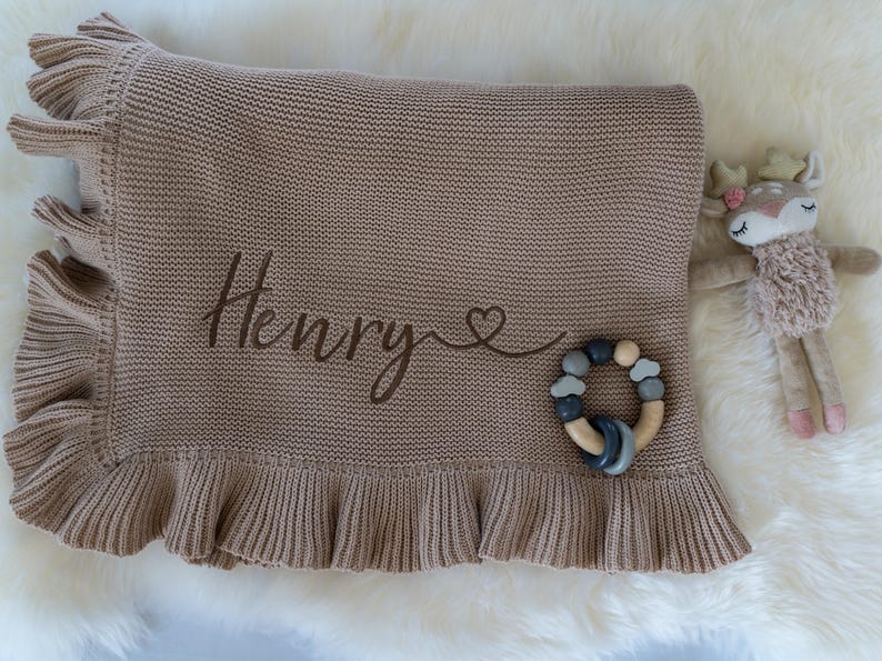 May include: A tan knitted baby blanket with a ruffled edge, embroidered with the name "Henry" and a heart. A wooden and silicone teether and a stuffed deer toy are next to the blanket.