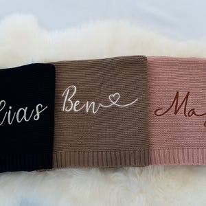 May include: Three personalized knitted blankets in black, brown, and pink. Each blanket features a name embroidered in a contrasting color: "Elias" in white, "Ben" with a heart in white, and "Maja" in brown. The blankets are displayed on a white, fluffy surface.