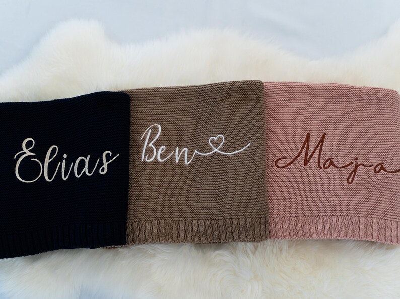 May include: Three personalized knitted blankets in black, brown, and pink. Each blanket features a name embroidered in a contrasting color: "Elias" in white, "Ben" with a heart in white, and "Maja" in brown. The blankets are displayed on a white, fluffy surface.