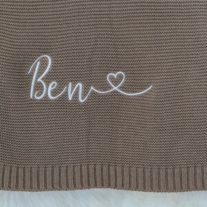 May include: A brown knitted blanket with the name "Ben" embroidered in white script, followed by a heart symbol. The blanket has a ribbed border and a soft, textured appearance, suitable for baby or child.
