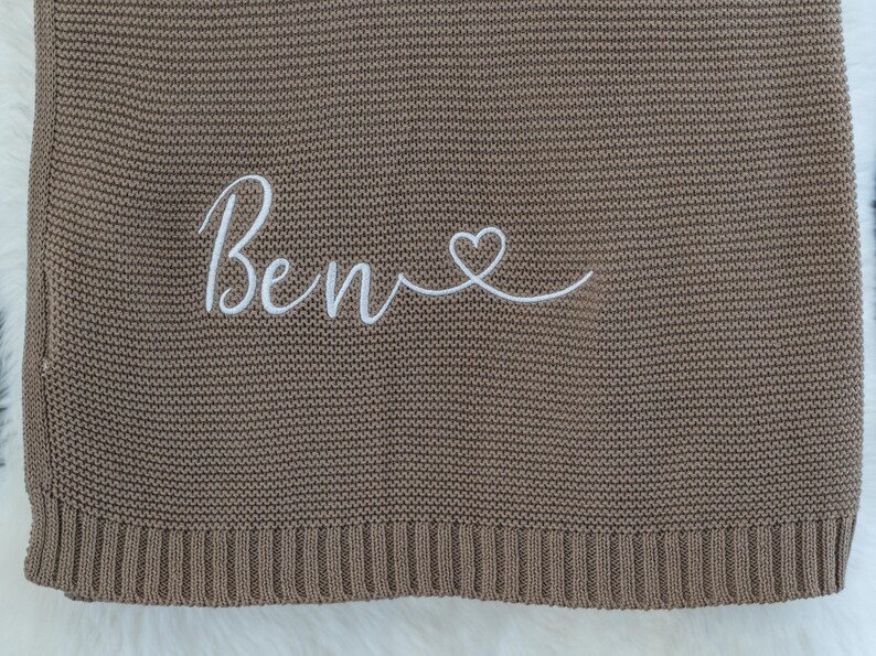 May include: A brown knitted blanket with the name "Ben" embroidered in white script, followed by a heart symbol. The blanket has a ribbed border and a soft, textured appearance, suitable for baby or child.