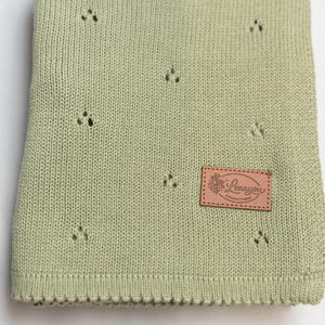 May include: A sage green knitted blanket featuring a repeating pattern of small, delicate eyelet holes. A rectangular tan leather-like patch is sewn onto the lower right corner, displaying the text Lenayou COLLECTIONS with a floral graphic.