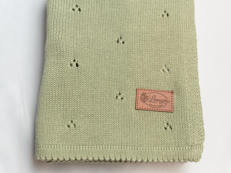 May include: A sage green knitted blanket featuring a repeating pattern of small, delicate eyelet holes. A rectangular tan leather-like patch is sewn onto the lower right corner, displaying the text Lenayou COLLECTIONS with a floral graphic.