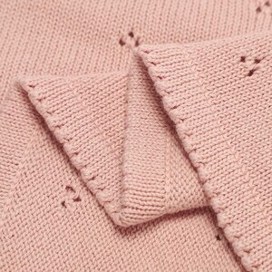 May include: A soft, light pink knitted fabric featuring a delicate, repeating eyelet pattern throughout. The material displays a consistent stockinette stitch texture with a finished, ribbed edge along the folded section of the textile.