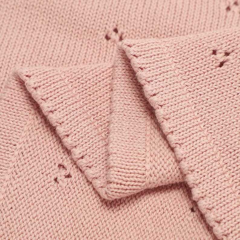 May include: A soft, light pink knitted fabric featuring a delicate, repeating eyelet pattern throughout. The material displays a consistent stockinette stitch texture with a finished, ribbed edge along the folded section of the textile.