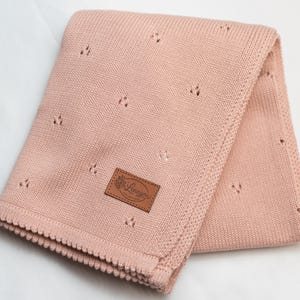 May include: A soft, dusty pink knitted blanket featuring a delicate pointelle pattern of small, repeating eyelet clusters. The blanket has a textured, scalloped border and a rectangular brown leather-like patch with the text Lenayou.