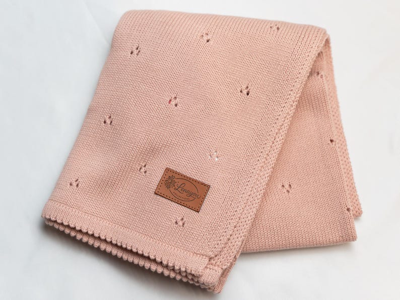May include: A soft, dusty pink knitted blanket featuring a delicate pointelle pattern of small, repeating eyelet clusters. The blanket has a textured, scalloped border and a rectangular brown leather-like patch with the text Lenayou.