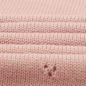 May include: A close-up view of a light pink knitted fabric featuring a consistent stockinette stitch pattern. The material displays horizontal folds that create depth and texture across the surface, with a small, singular snag visible.