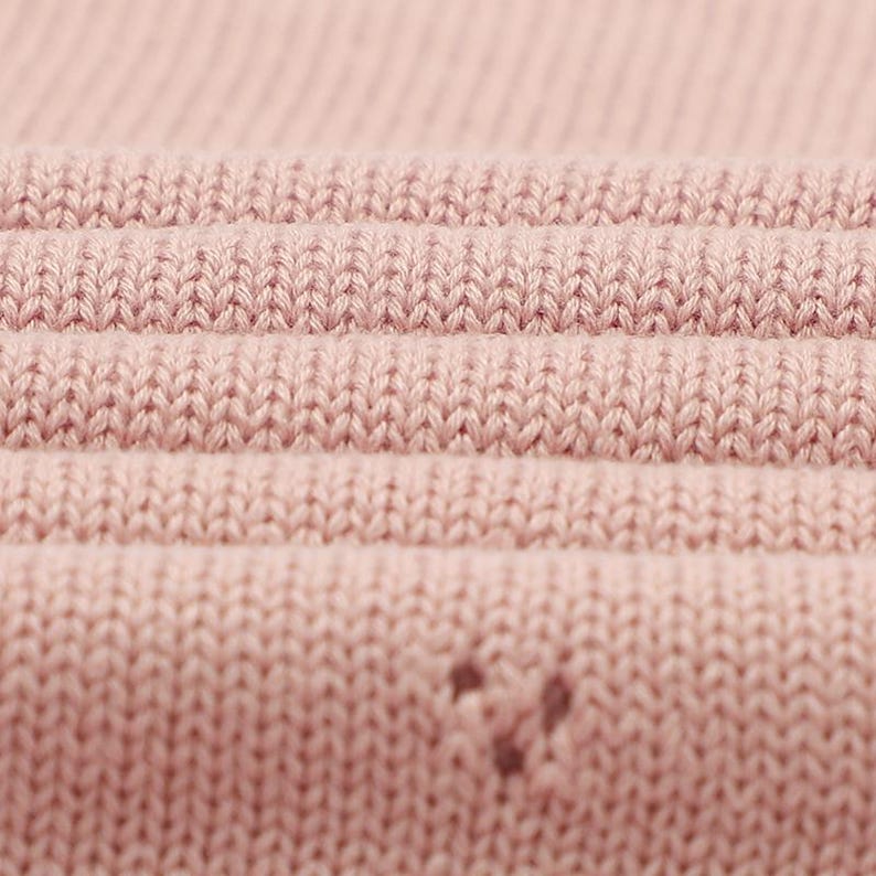 May include: A close-up view of a light pink knitted fabric featuring a consistent stockinette stitch pattern. The material displays horizontal folds that create depth and texture across the surface, with a small, singular snag visible.
