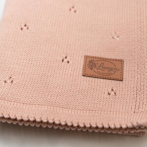 May include: A dusty pink knitted blanket featuring a subtle pointelle pattern of small triangular cutouts. A rectangular brown faux leather patch is stitched onto the corner, displaying the text Lenayou Collections with a floral graphic.