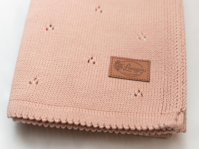 May include: A dusty pink knitted blanket featuring a subtle pointelle pattern of small triangular cutouts. A rectangular brown faux leather patch is stitched onto the corner, displaying the text Lenayou Collections with a floral graphic.