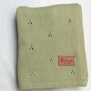 May include: A sage green knitted blanket featuring a repeating pattern of small, three-hole eyelet clusters. A rectangular brown leather-like patch is stitched onto the lower right corner, displaying the text Lenayou Collections in script.