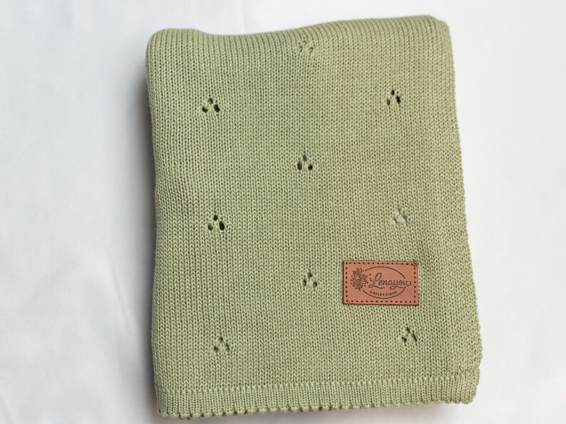 May include: A sage green knitted blanket featuring a repeating pattern of small, three-hole eyelet clusters. A rectangular brown leather-like patch is stitched onto the lower right corner, displaying the text Lenayou Collections in script.