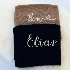 May include: Two knitted blankets, one tan and one navy blue, are stacked on a white fluffy surface. The tan blanket has the name "Bene" embroidered in white script with a heart. The navy blanket has the name "Elias" embroidered in white script.