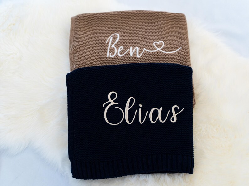 May include: Two knitted blankets, one tan and one navy blue, are stacked on a white fluffy surface. The tan blanket has the name "Bene" embroidered in white script with a heart. The navy blanket has the name "Elias" embroidered in white script.