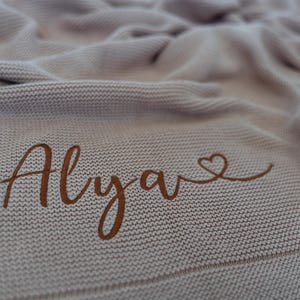 May include: A light brown knitted blanket with the name "Alya" in a cursive font, accompanied by a heart symbol. The blanket has a textured appearance and is folded, creating soft shadows.