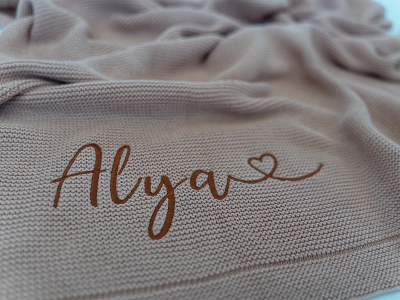 May include: A light brown knitted blanket with the name "Alya" in a cursive font, accompanied by a heart symbol. The blanket has a textured appearance and is folded, creating soft shadows.
