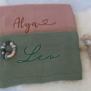May include: Two personalized baby blankets, one pink and one green, with names embroidered in cursive. A wooden and silicone teething ring and a plush deer toy are also visible. The pink blanket has the name "Alya" and the green blanket has the name "Leo".
