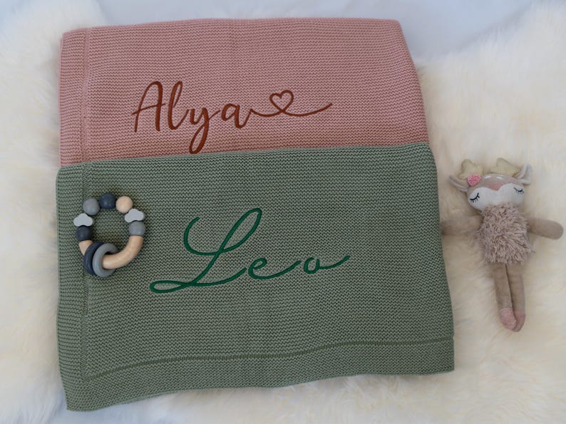 May include: Two personalized baby blankets, one pink and one green, with names embroidered in cursive. A wooden and silicone teething ring and a plush deer toy are also visible. The pink blanket has the name "Alya" and the green blanket has the name "Leo".