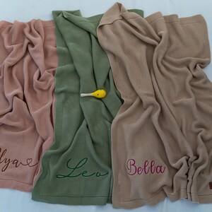 May include: Three soft, ribbed knit blankets in shades of pink, green, and tan. Each blanket is personalized with a name embroidered in a complementary color. A small yellow rattle rests on the green blanket.