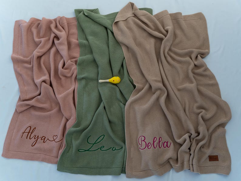 May include: Three soft, ribbed knit blankets in shades of pink, green, and tan. Each blanket is personalized with a name embroidered in a complementary color. A small yellow rattle rests on the green blanket.