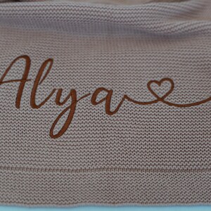 May include: A knitted blanket in a dusty rose color. The blanket features the name "Alyave" embroidered in a darker brown thread, with a heart symbol at the end. The blanket has a textured appearance.