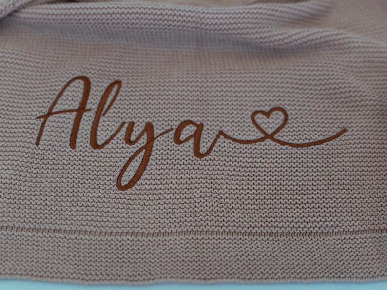 May include: A knitted blanket in a dusty rose color. The blanket features the name "Alyave" embroidered in a darker brown thread, with a heart symbol at the end. The blanket has a textured appearance.