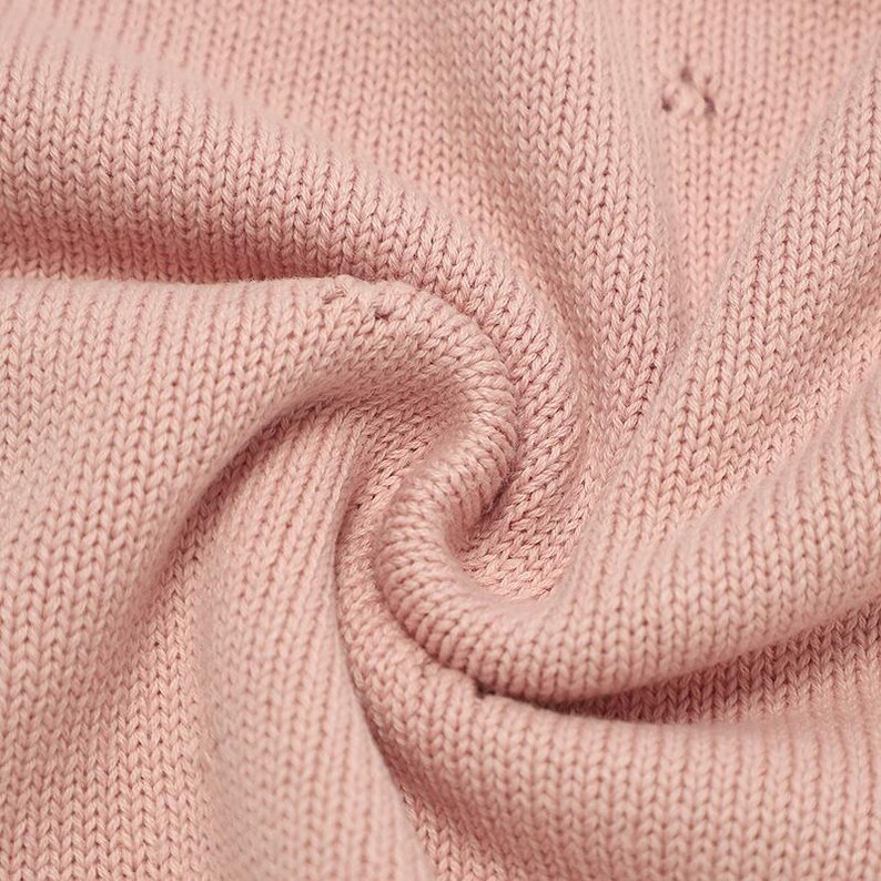 May include: A close-up view of a soft, light pink knitted fabric with a consistent stockinette stitch pattern. The material is gathered into a gentle swirl, highlighting the texture of the yarn loops and the uniform, matte finish of the textile.