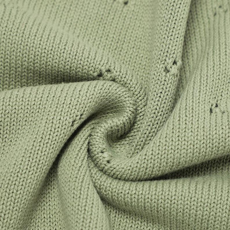 May include: A sage green knit fabric featuring a consistent stockinette stitch pattern with small, evenly spaced eyelet holes integrated into the weave. The material is folded into soft, deep pleats, highlighting the texture of the yarn.