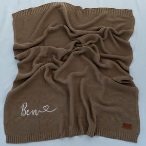 May include: A brown knitted blanket with the name "Ben" embroidered in white script with a heart symbol. The blanket is folded and draped, showing a textured weave. A small brown label is sewn on the corner.