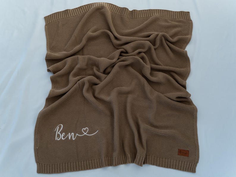 May include: A brown knitted blanket with the name "Ben" embroidered in white script with a heart symbol. The blanket is folded and draped, showing a textured weave. A small brown label is sewn on the corner.