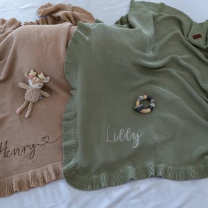 May include: Two soft, ruffled baby blankets in tan and sage green, personalized with names. A stuffed deer toy and a wooden teething ring rest on the blankets. The blanket in tan is embroidered with the name "Henry". The green blanket is embroidered with the name "Lilly".