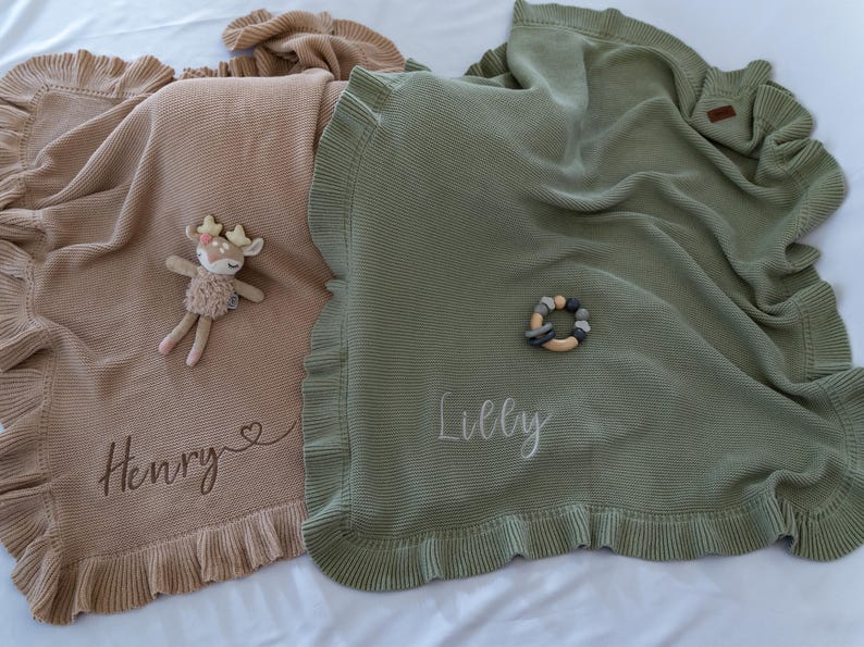 May include: Two soft, ruffled baby blankets in tan and sage green, personalized with names. A stuffed deer toy and a wooden teething ring rest on the blankets. The blanket in tan is embroidered with the name "Henry". The green blanket is embroidered with the name "Lilly".